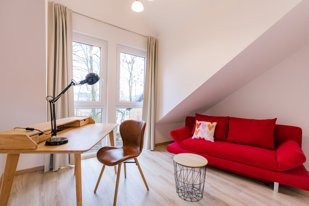 Gemütliche Arbeitsecke mit Holzschreibtisch und rotem Schlafsofa unter Dachschräge