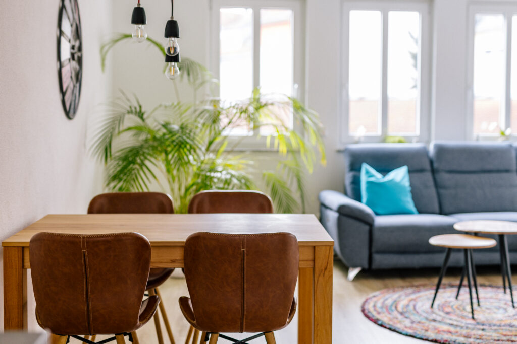 Esstisch mit Lederstühlen, dahinter graues Sofa und helle Fenster
