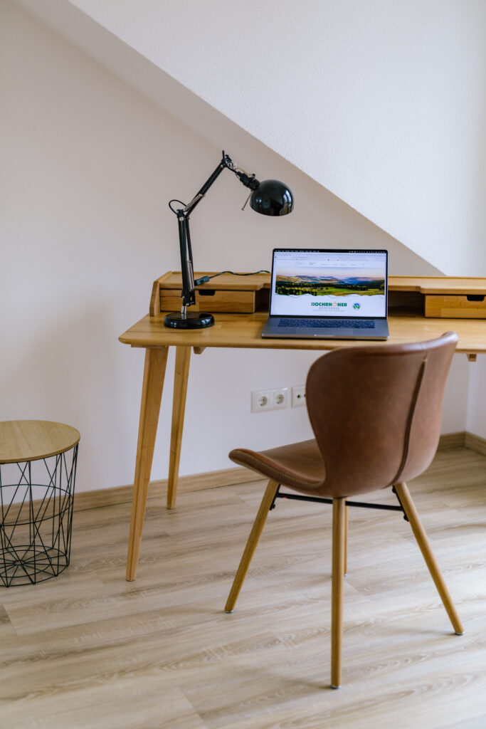 Holzschreibtisch mit Laptop und Arbeitsplatzlampe, dazu brauner Lederstuhl unter Dachschräge