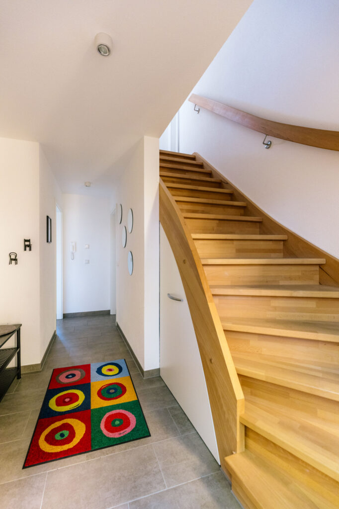 Heller Eingangsbereich des Ferienhauses mit geschwungener Holztreppe und buntem Teppich